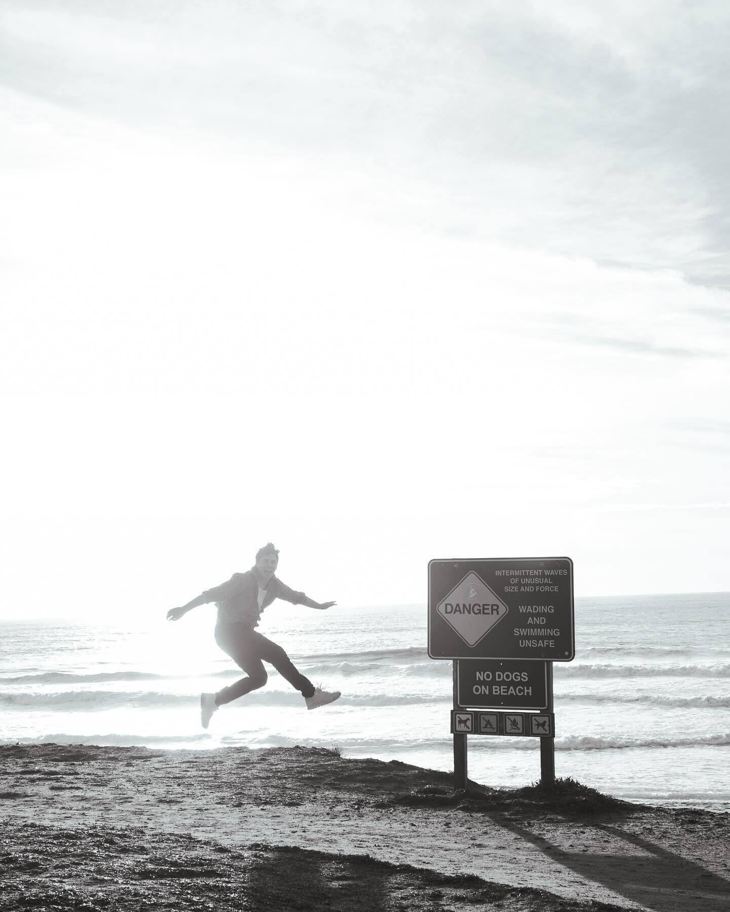 A figure leaps beside a warning sign on a sea cliff, backlit, in black and white
