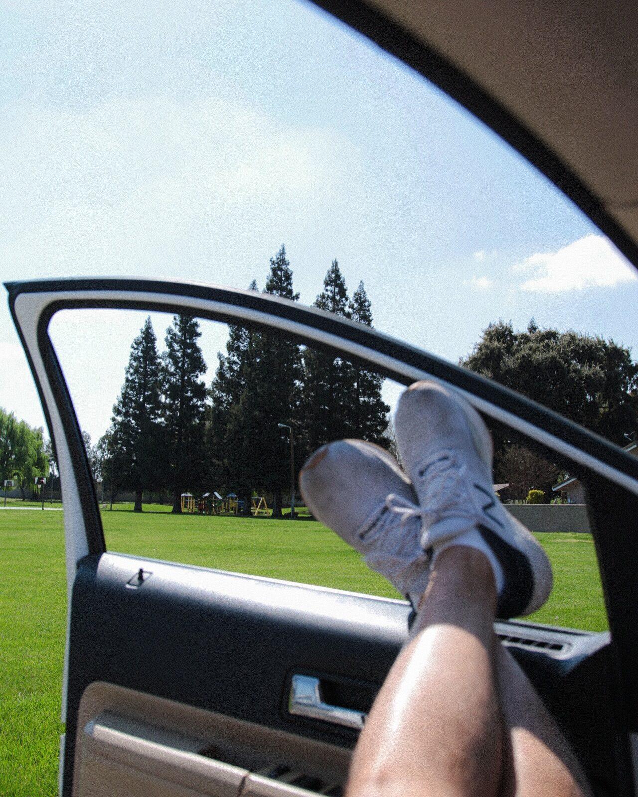Sneakers propped on an open car door, pines and a green field beyond
