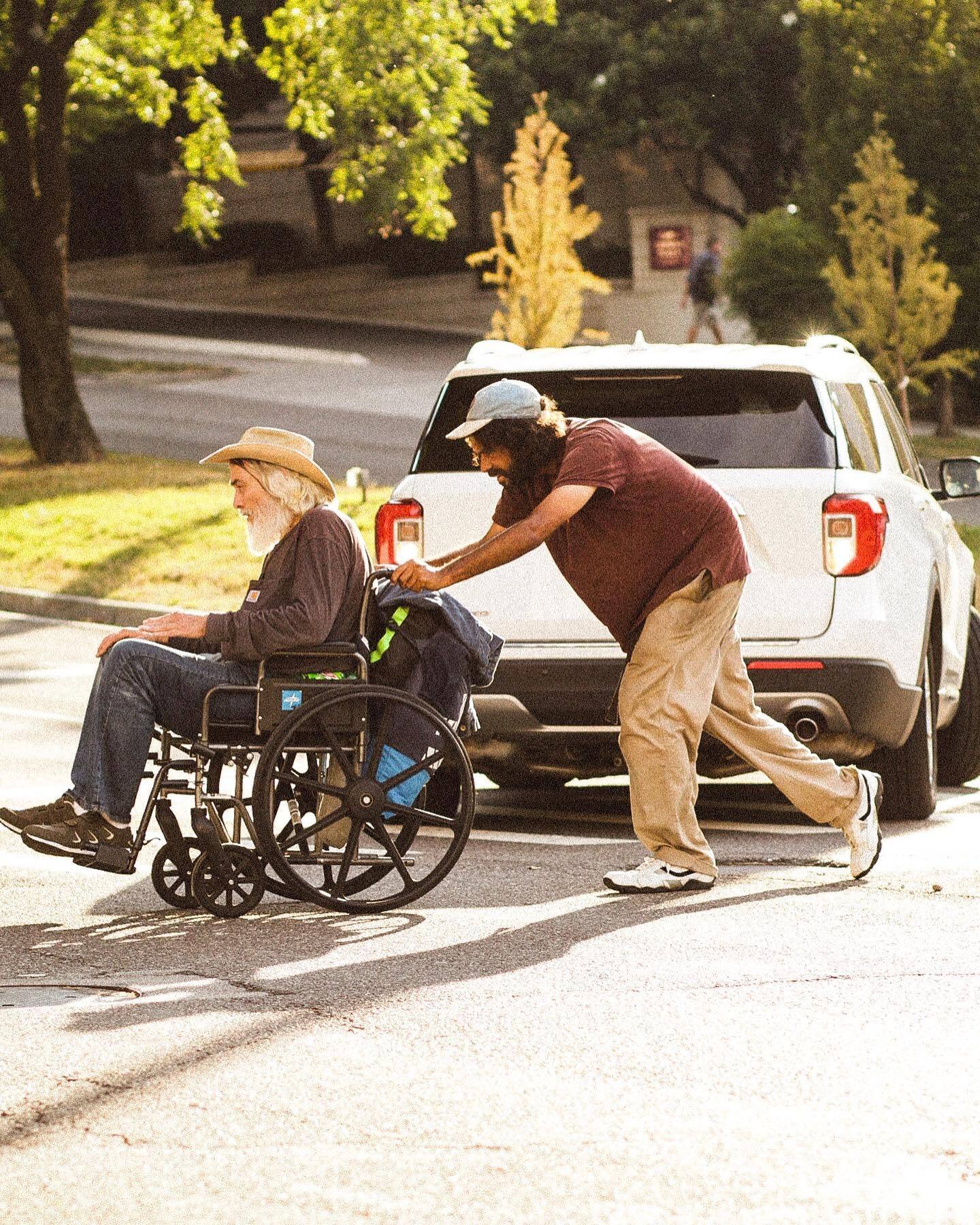 A man in a straw hat pushes another man in a wheelchair past a white pickup