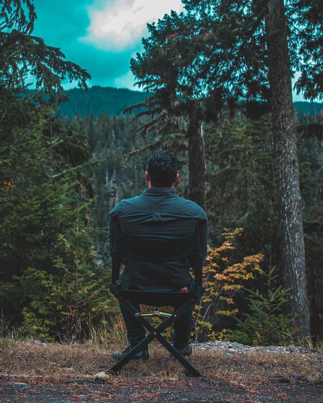 A man in a camp chair faces a wall of pines
