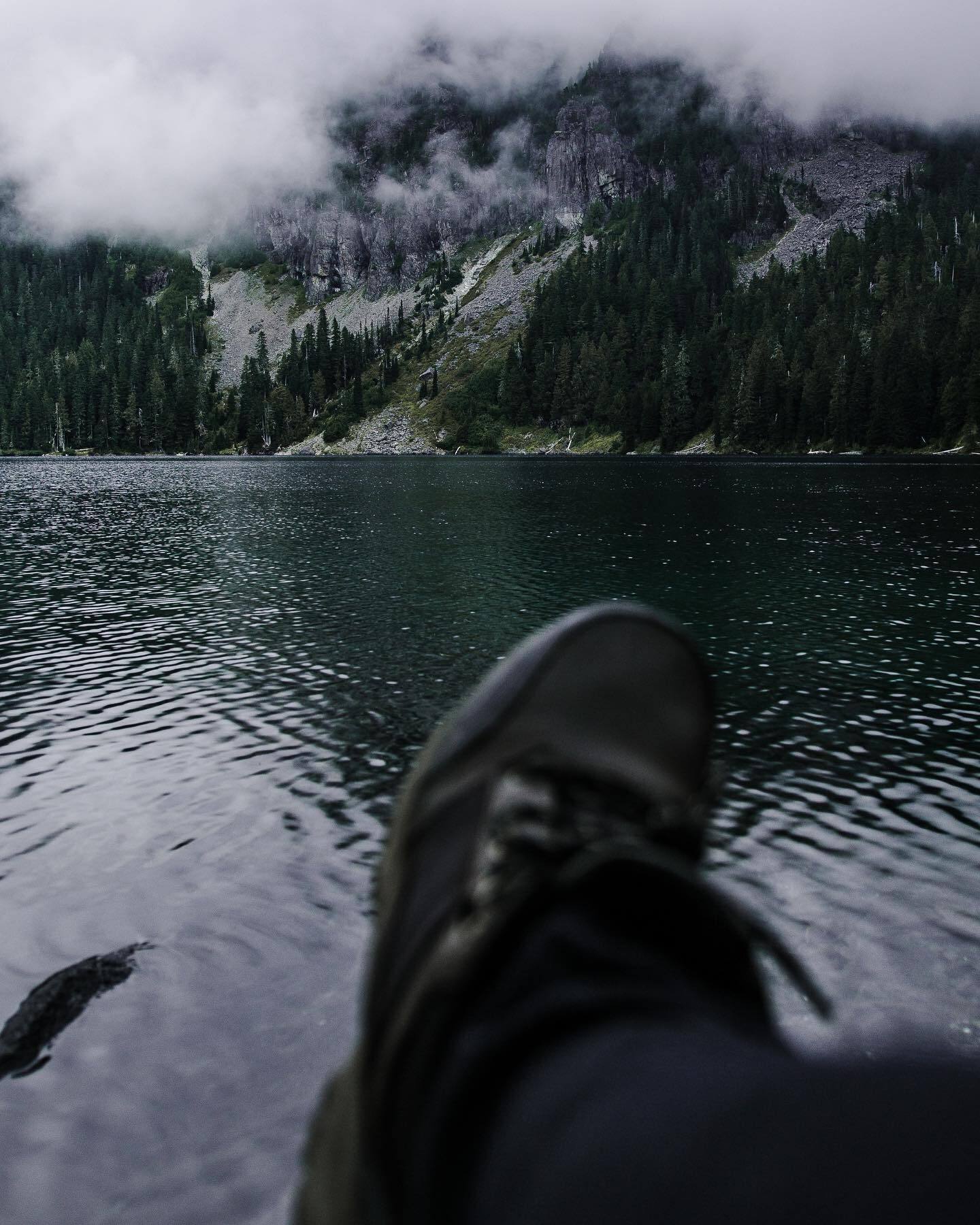 A boot held out over a still lake beneath cloud-covered peaks